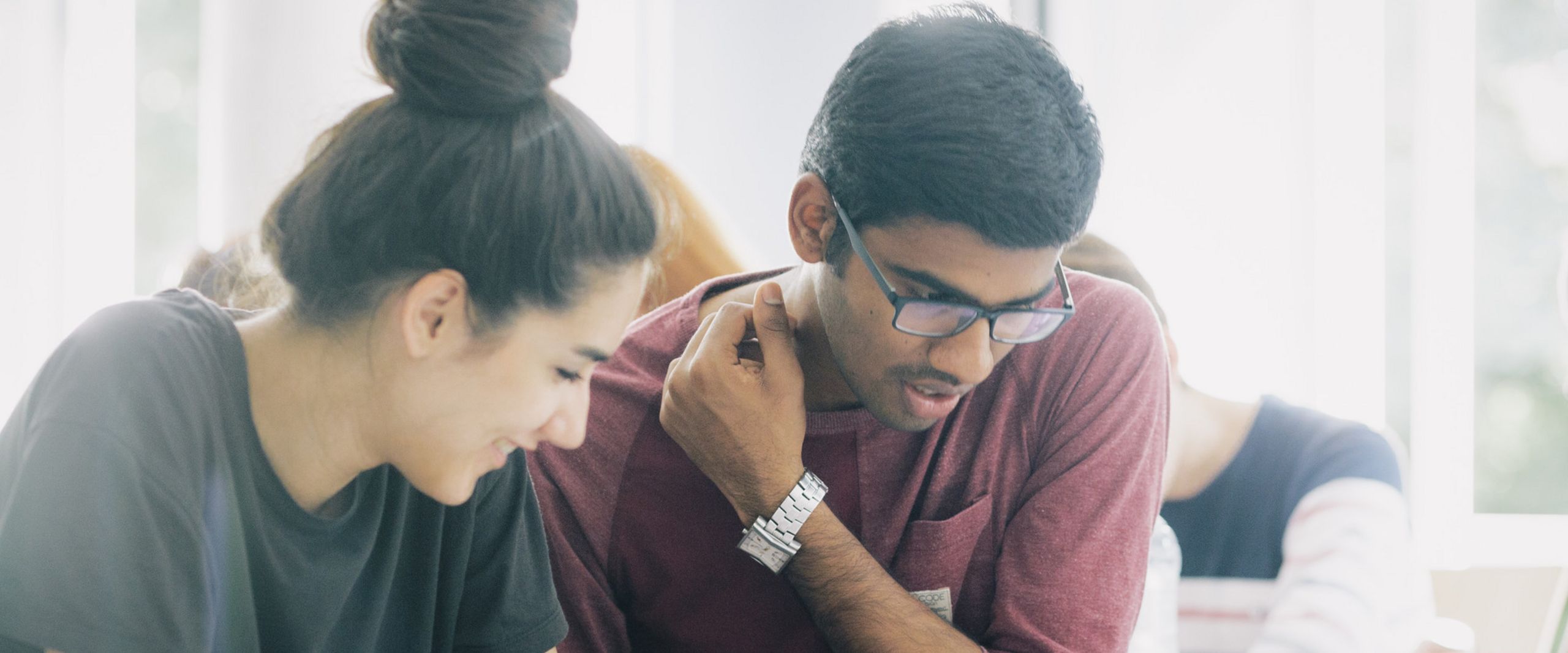 Eine Studentin und ein Student sitzen nebeneinander und blicken gemeinsam auf Unterlagen vor sich auf dem Tisch.