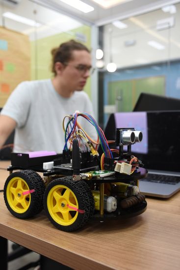 Student sitzt hinter einem Kleinroboter mit Laptop
