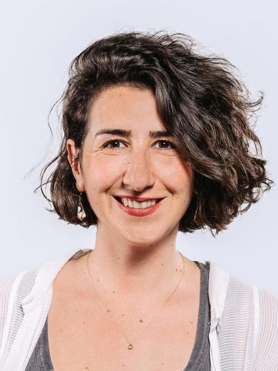 Auf dem Bild ist Duygu Rengail-Wenck zusehen. Sie hat kinnlanges braunes, lockiges Haar. Sie trägt eine offene weiße Bluse über einem grünen Top. 