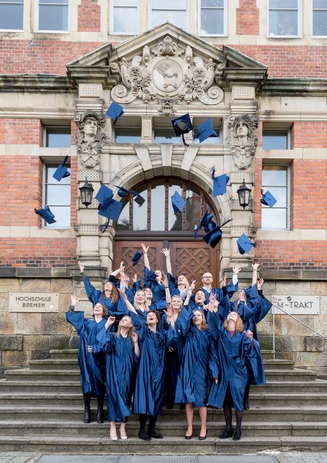 HSB Absolvent:innen stehen in Robe vor dem Gebäude und werfen ihre Hüte in die Luft.