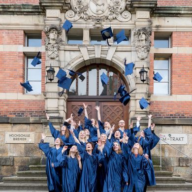 HSB Absolvent:innen stehen in Robe vor dem Gebäude und werfen ihre Hüte in die Luft.