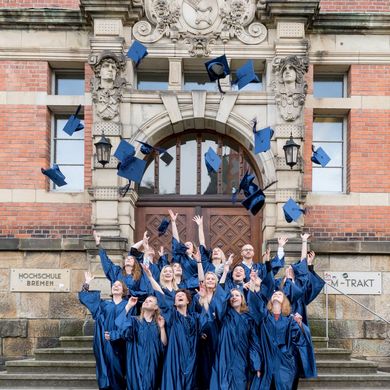 HSB Absolvent:innen stehen in Robe vor dem Gebäude und werfen ihre Hüte in die Luft.