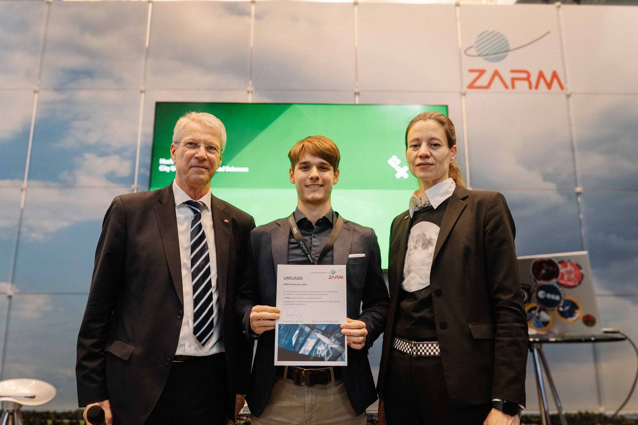 Gruppenbild mit zwei Männern und einer Frau. Der Mann in der Mitte hält eine Urkunde.