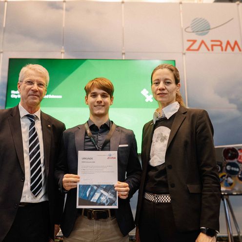 Gruppenbild mit zwei Männern und einer Frau. Der Mann in der Mitte hält eine Urkunde.