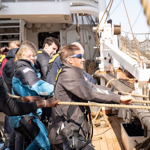 Auf dem Bild sind Leute zu erkennen, die an Board eines Schiffes an Seilen ziehen. 