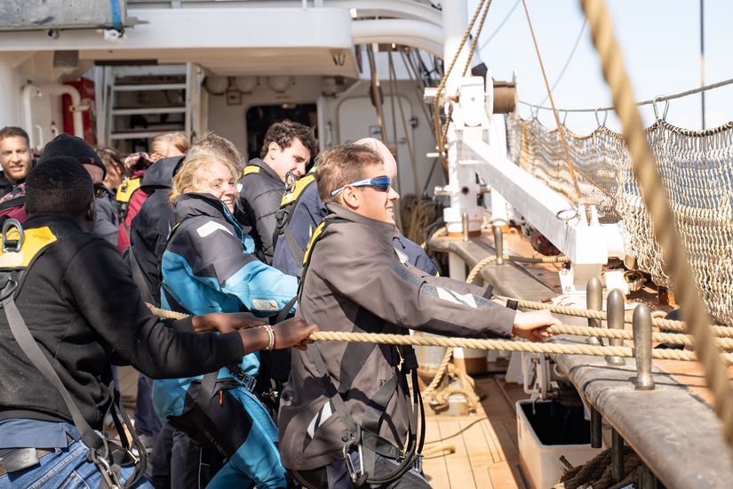 Auf dem Bild sind Leute zu erkennen, die an Board eines Schiffes an Seilen ziehen. 
