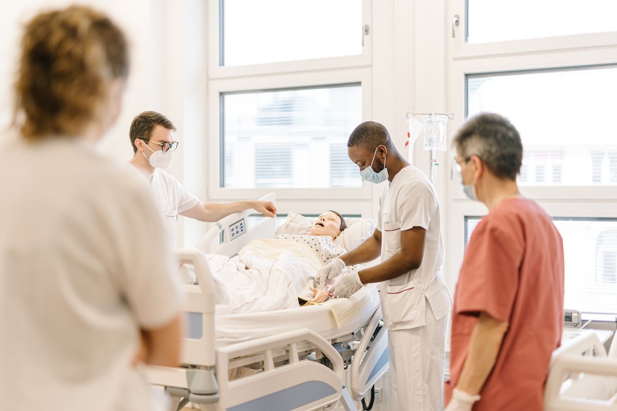 Pflegestudierende &uuml;ben an einem Krankenhausbett mit einer Puppe im Simulationszentrum des Studiengangs.