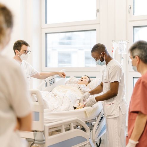 Pflegestudierende &uuml;ben an einem Krankenhausbett mit einer Puppe im Simulationszentrum des Studiengangs.
