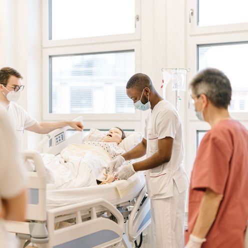 Pflegestudierende &uuml;ben an einem Krankenhausbett mit einer Puppe im Simulationszentrum des Studiengangs.