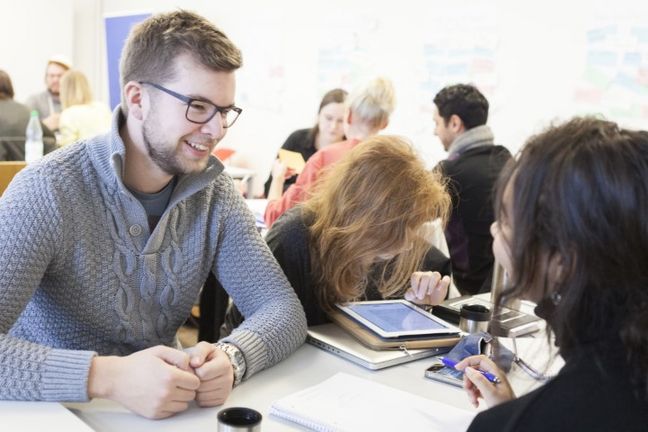 Bild zeigt einen junge, freundlich schauenden Studenten neben Studentinnen an einem Tisch