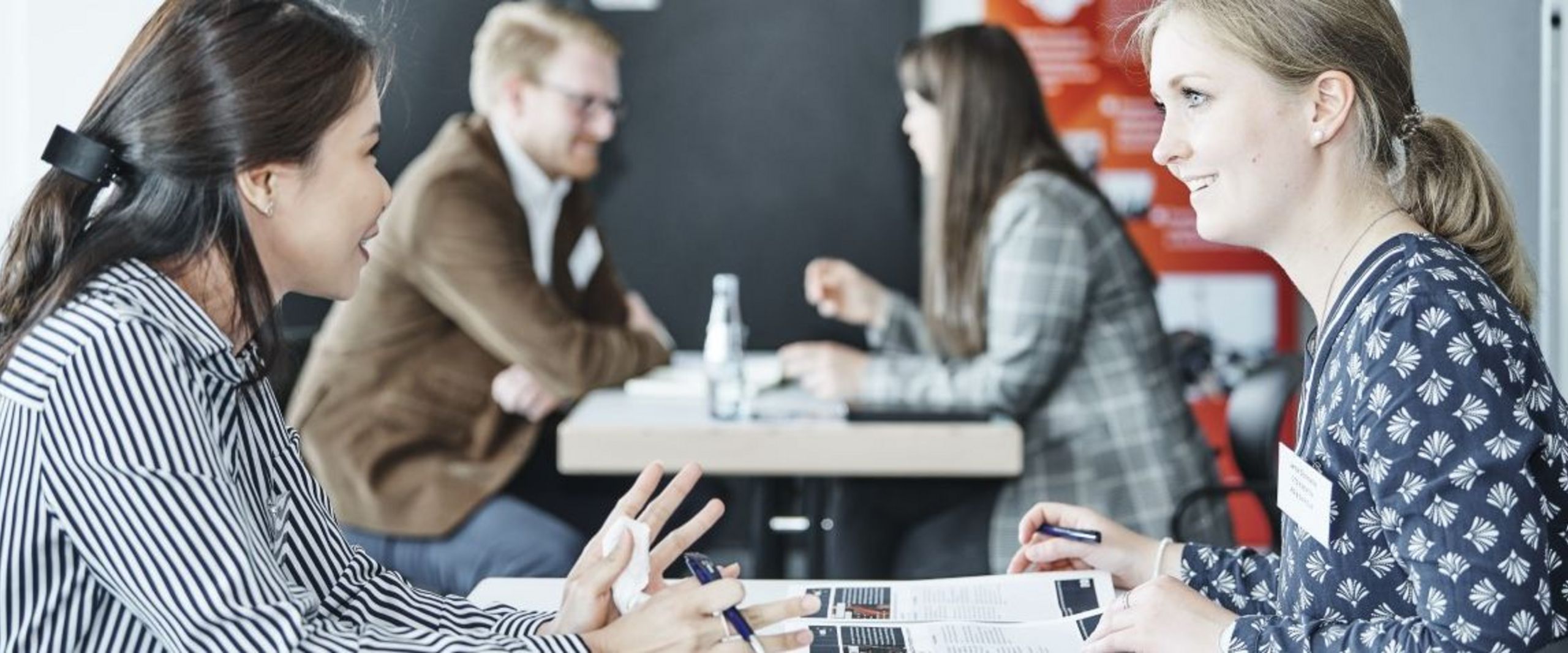 Männer und Frauen sitzen zu zweit an einem Tisch gegenüber und unterhalten sich.