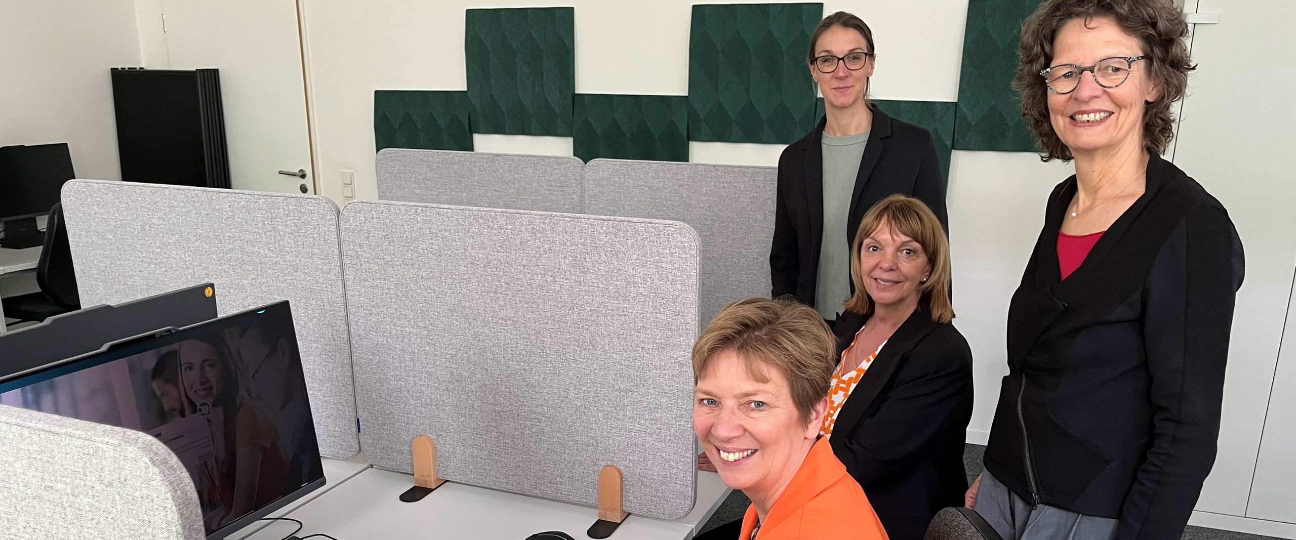 Vier Frauen sitzen und stehen an Tischen mit Bildschirmen und lächeln in die Kamera.
