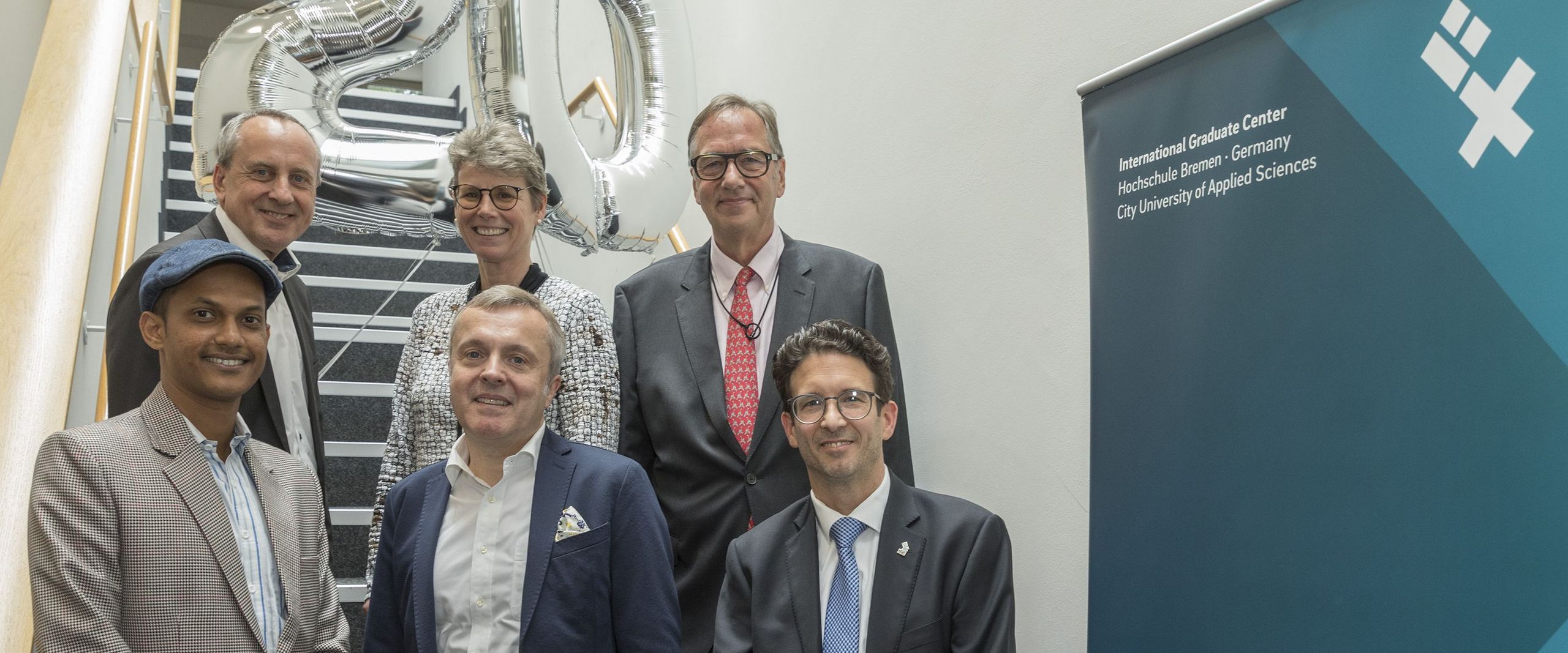 gruppenbild mit Männern und Frauen auf einer Treppe. Hinter ihnen schwebt eine 20 als zwei silberne Luftballons.