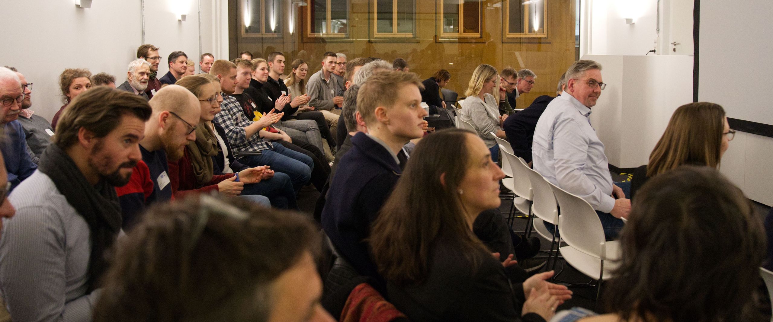 Das Bild zeigt einen Raum mit Zuhörer:innen, die auf Stühlen sitzen.