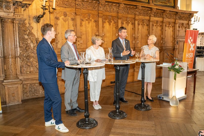 Five men and women stand at bar tables and discuss.