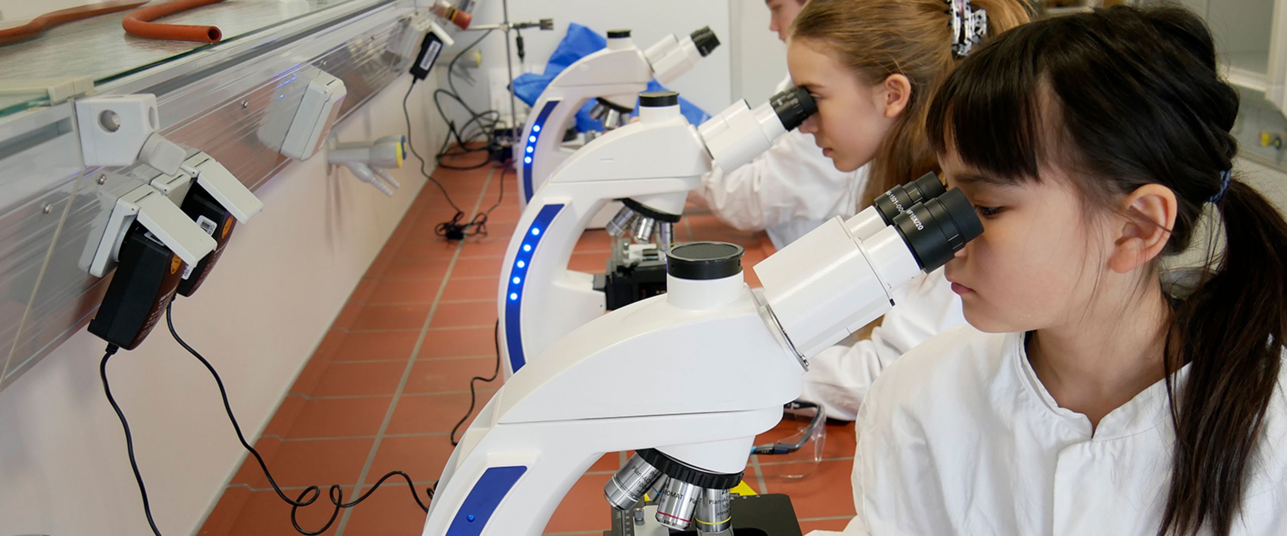 Drei M&auml;dchen sitzen an einem Tisch in einem Labor und bauen etwas mit kleinen R&auml;dern.