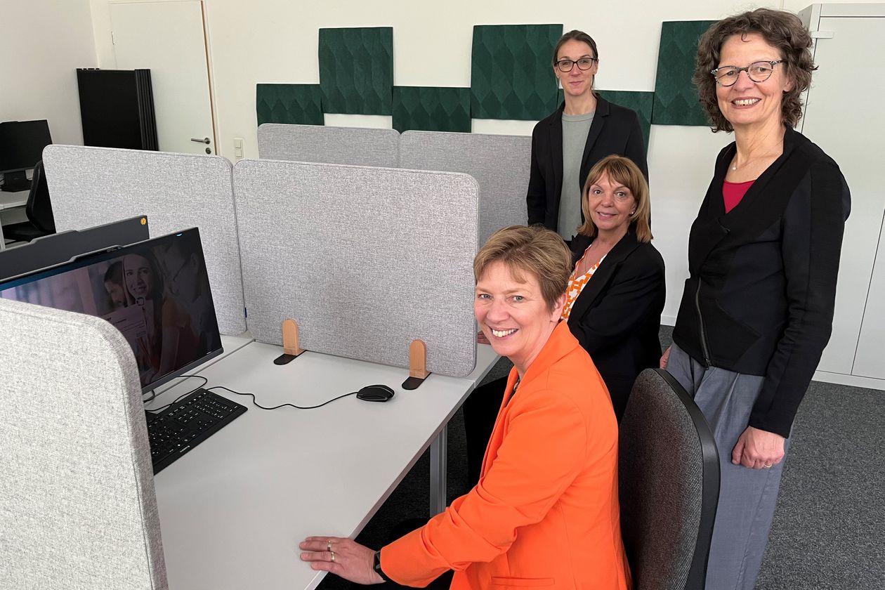 Vier Frauen sitzen und stehen an Tischen mit Bildschirmen und l&auml;cheln in die Kamera.
