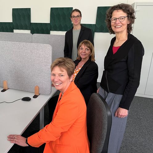 Vier Frauen sitzen und stehen an Tischen mit Bildschirmen und l&auml;cheln in die Kamera.