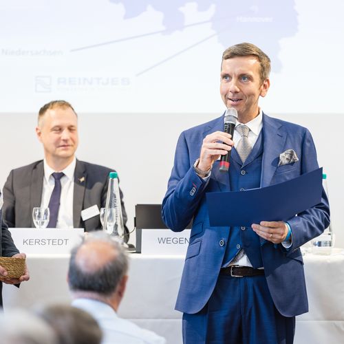 Mann im Anzug spricht in Mikro. Links h&ouml;ren ihm Menschen zu
