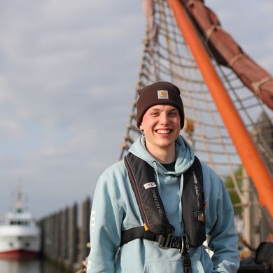 Junger Mann mit Schwimmweste, Pullover und M&uuml;tze auf einem Schiff.