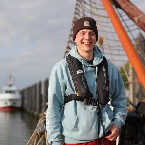 Junger Mann mit Schwimmweste, Pullover und M&uuml;tze auf einem Schiff.
