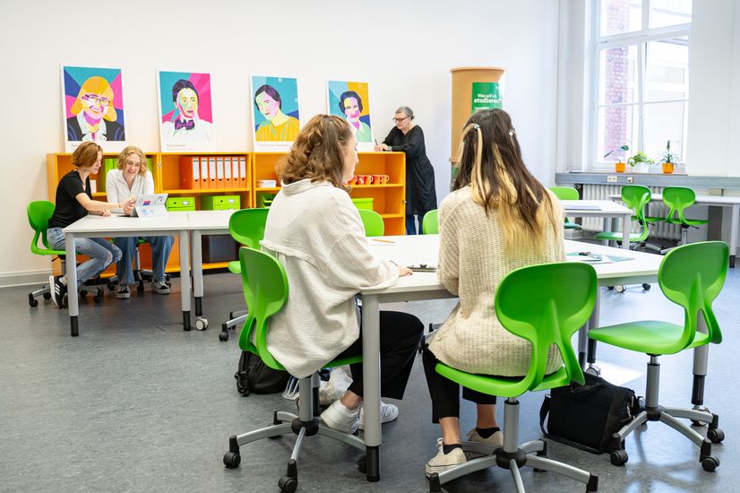 Raum mit Stühlen und Tischen. Frauen arbeiten daran. Eine Frau lehnt am Bücherregal.