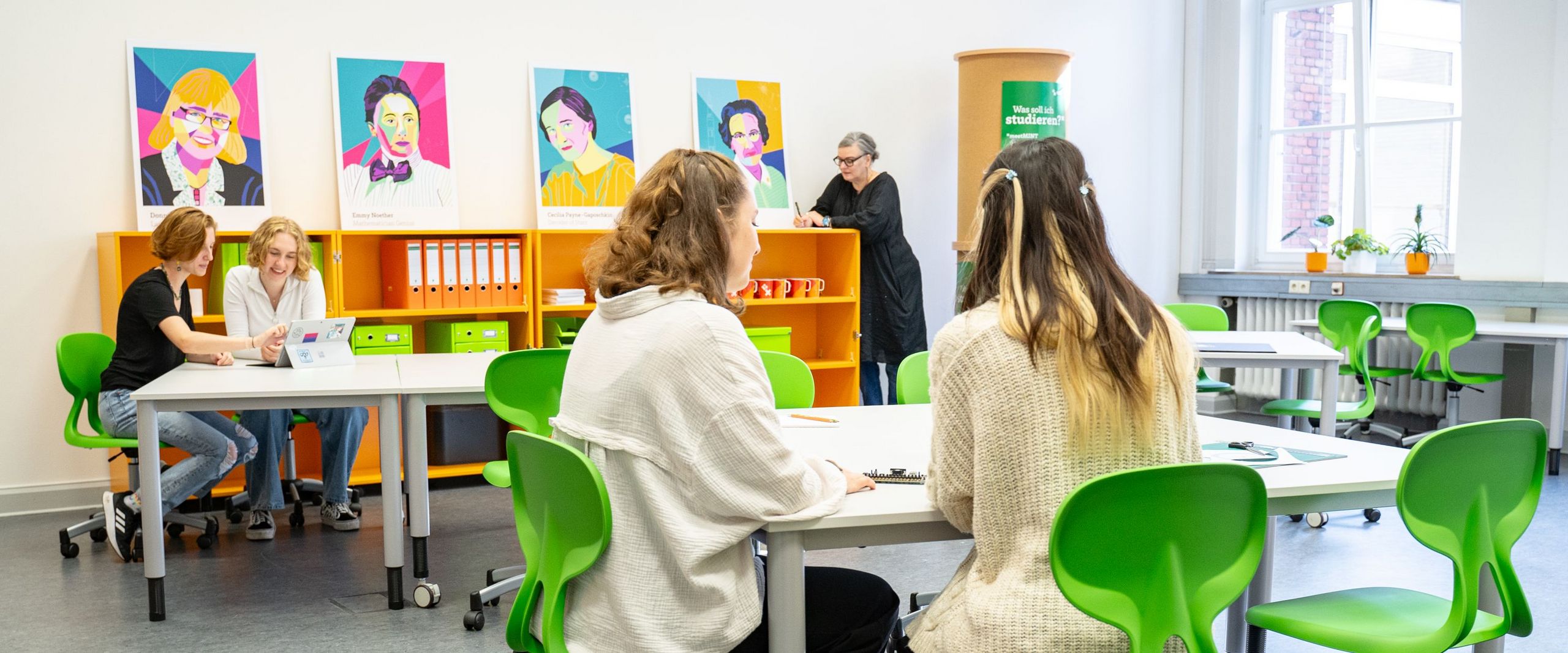 Raum mit Stühlen und Tischen. Frauen arbeiten daran. Eine Frau lehnt am Bücherregal.