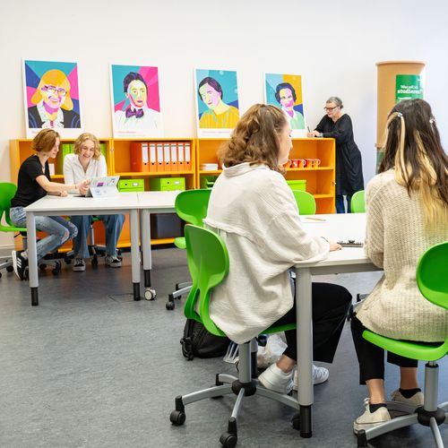Room with chairs and tables. Women are working at them. A woman leans against the bookshelf.