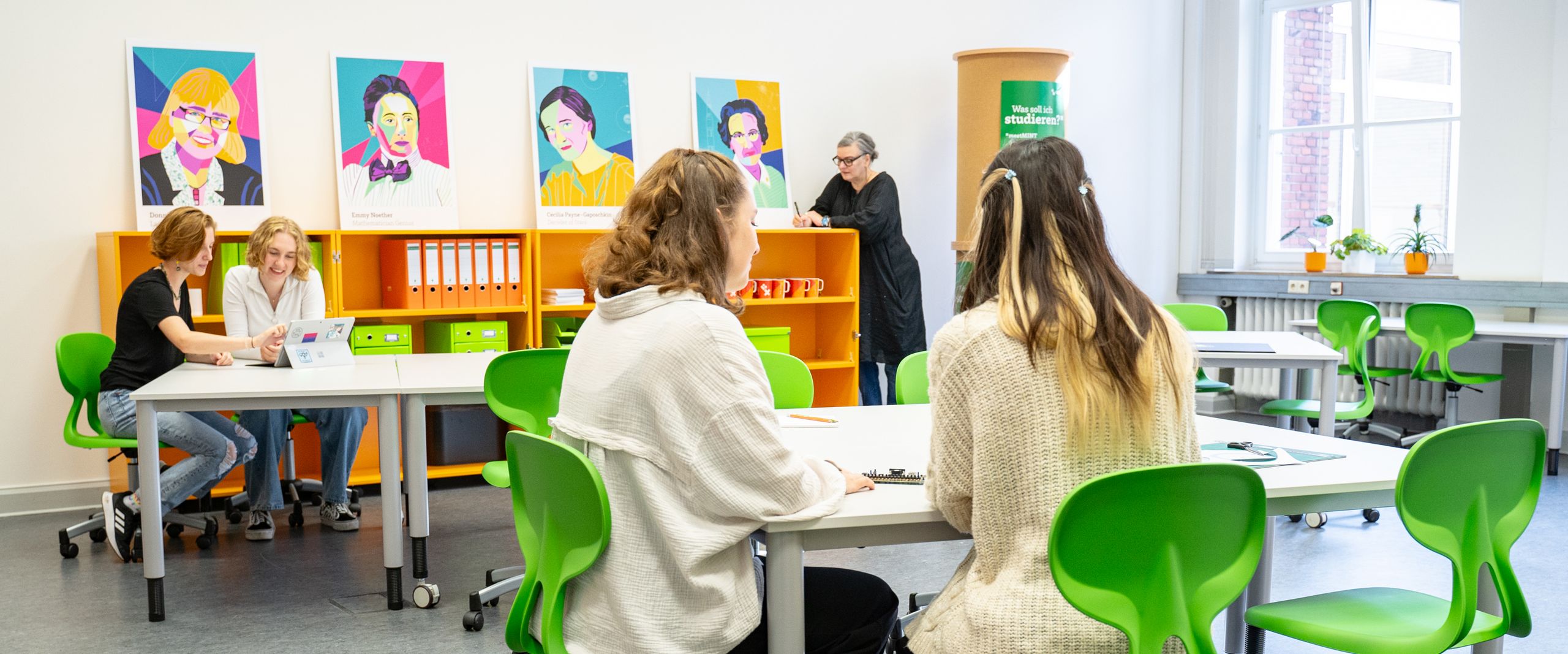 meetMINT-Space Räume Raum mit Stühlen und Tischen. Frauen arbeiten daran. Eine Frau lehnt am Bücherregal.