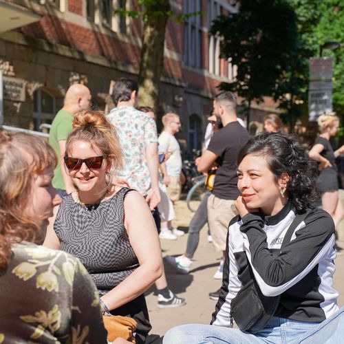 Drei Studentinnen sitzen bei sonnigem Wetter auf einer Bank vorm M-Geb&auml;ude am Campus Neustadt und reden miteinander. Im Hintergrund steht eine Gruppe Studierender und unterh&auml;lt sich ebenso.