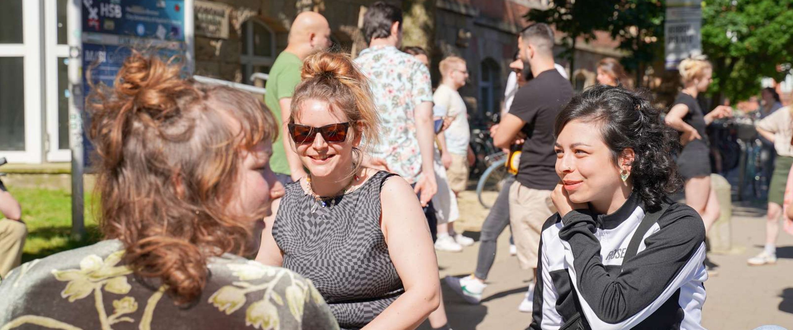 Studentinnen vorm M-Gebäude am Campus Neustadt Drei Studentinnen sitzen bei sonnigem Wetter auf einer Bank vorm M-Gebäude am Campus Neustadt und reden miteinander. Im Hintergrund steht eine Gruppe Studierender und unterhält sich ebenso.