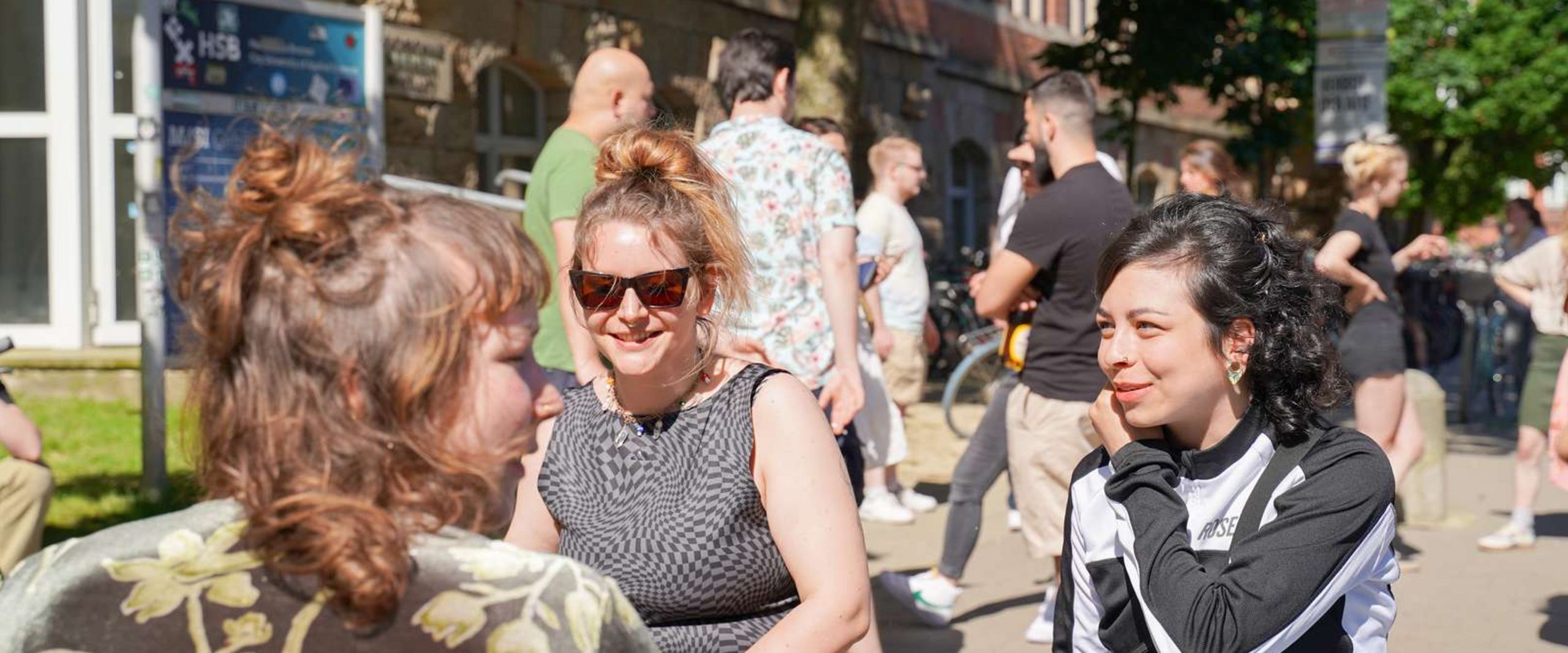 Drei Studentinnen sitzen bei sonnigem Wetter auf einer Bank vorm M-Geb&auml;ude am Campus Neustadt und reden miteinander. Im Hintergrund steht eine Gruppe Studierender und unterh&auml;lt sich ebenso.
