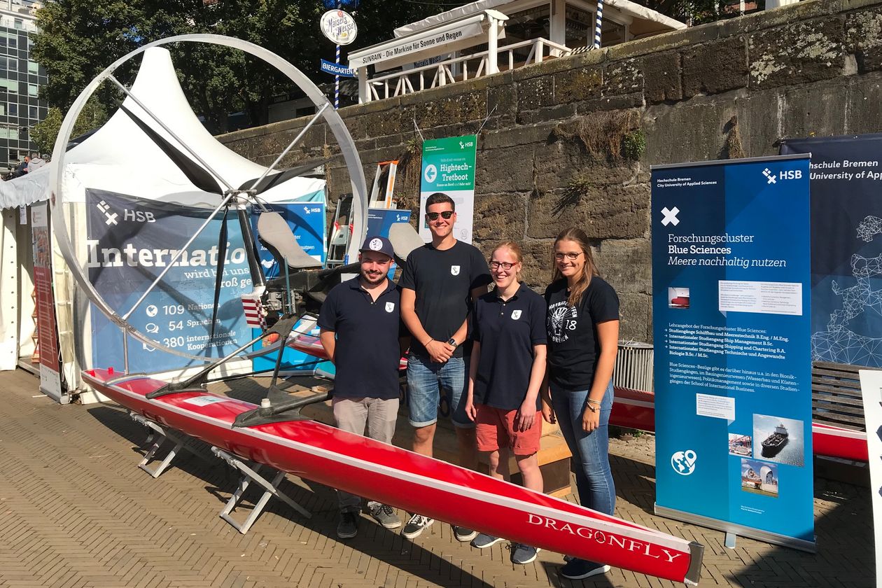 Junge Menschen stehen vor einem roten Tretboot. Rechts steht ein Banner und hinter ihnen eine weiße Pagode.