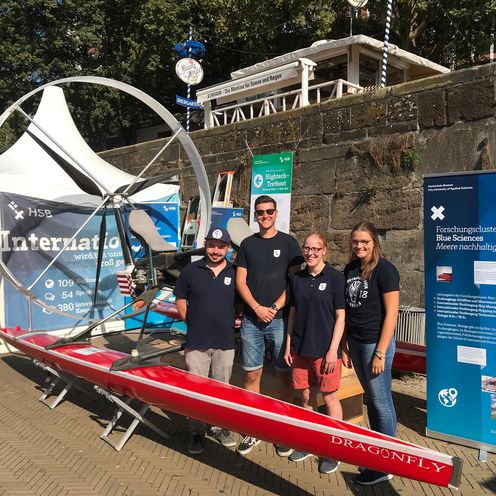 Junge Menschen stehen vor einem roten Tretboot. Rechts steht ein Banner und hinter ihnen eine wei&szlig;e Pagode.