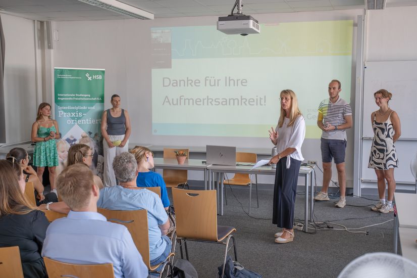 Studierende stehen neben Professorin Renate Freericks. Neben Ihnen steht ein Beamer und ein Roll-Up.