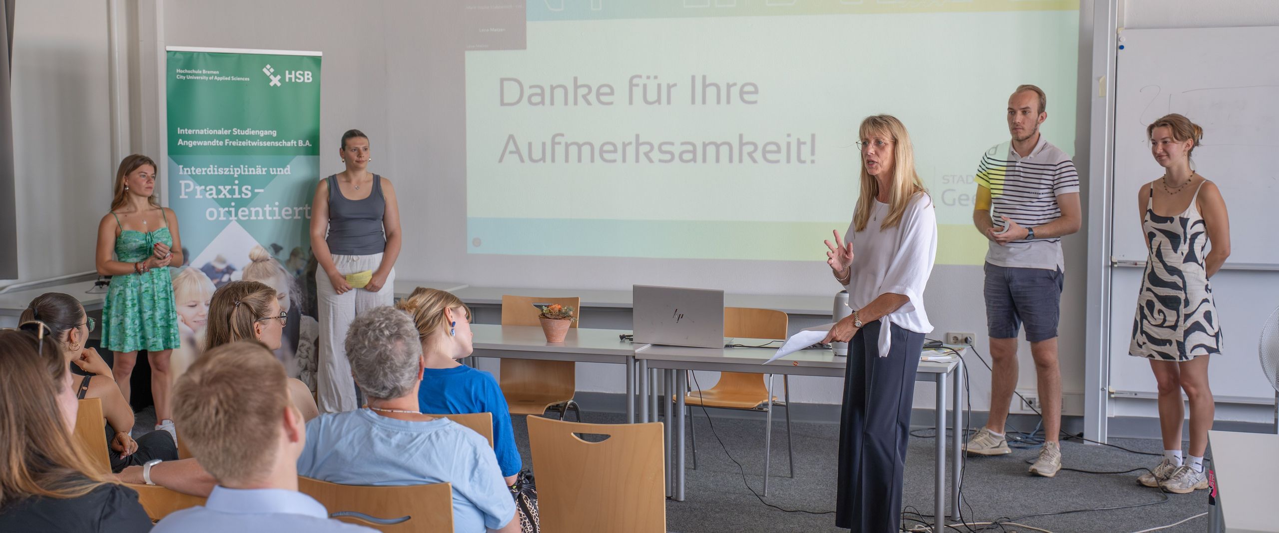 Studierende stehen neben Professorin Renate Freericks. Neben Ihnen steht ein Beamer und ein Roll-Up.