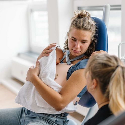 Auf dem Bild ist eine Studentin des Studiengangs Hebammen zu sehen, die eine Situation kurz nach der Geburt simuliert. Sie sitzt auf einem Stuhl und h&auml;lt eine Puppe im Arm, die ein Neugeborenes simuliert.