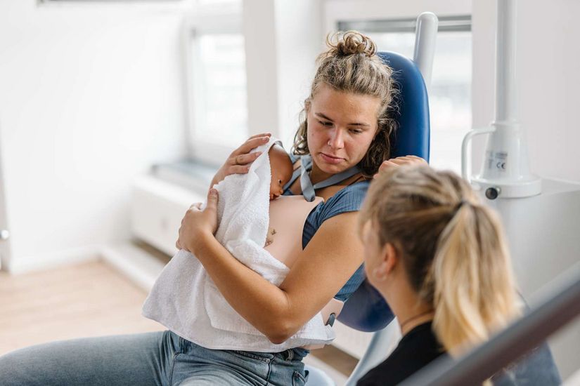 Studiengang Hebammen The picture shows a student on the midwifery degree programme simulating a situation shortly after giving birth. She is sitting on a chair and holding a doll in her arms that simulates a newborn baby.
