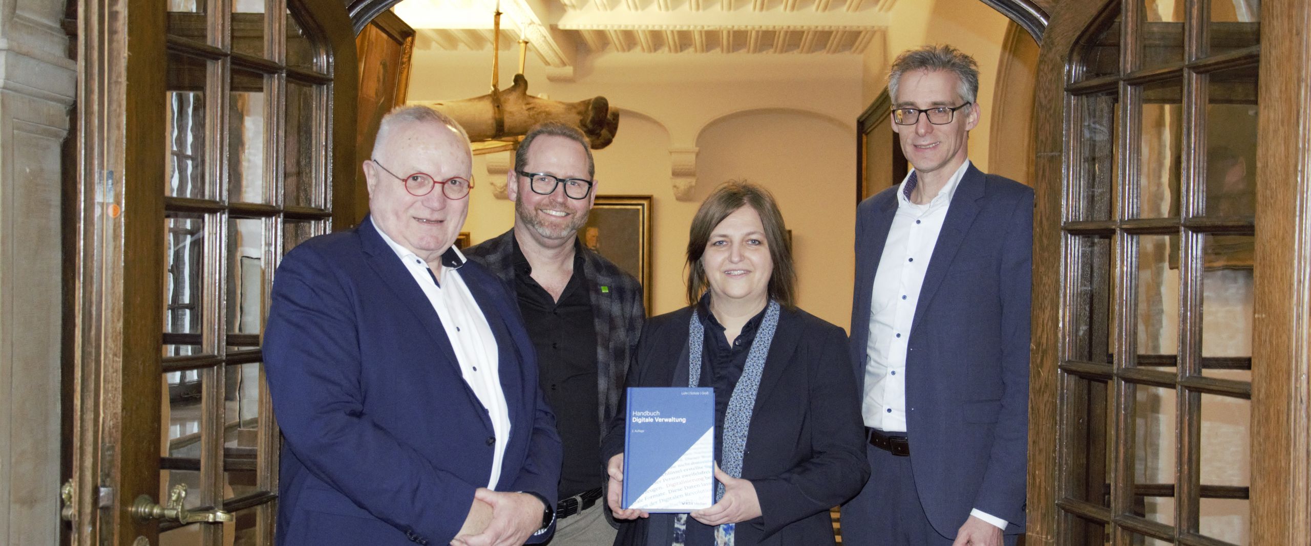 Gruppenbild mit drei Männern und einer Frau. Diese präsentiert in ihren Händen ein Buch. Alle stehen im Durchgang einer repräsentativen Holztür mit Glasfenstern. Beide Türflügel sind weit geöffnet.