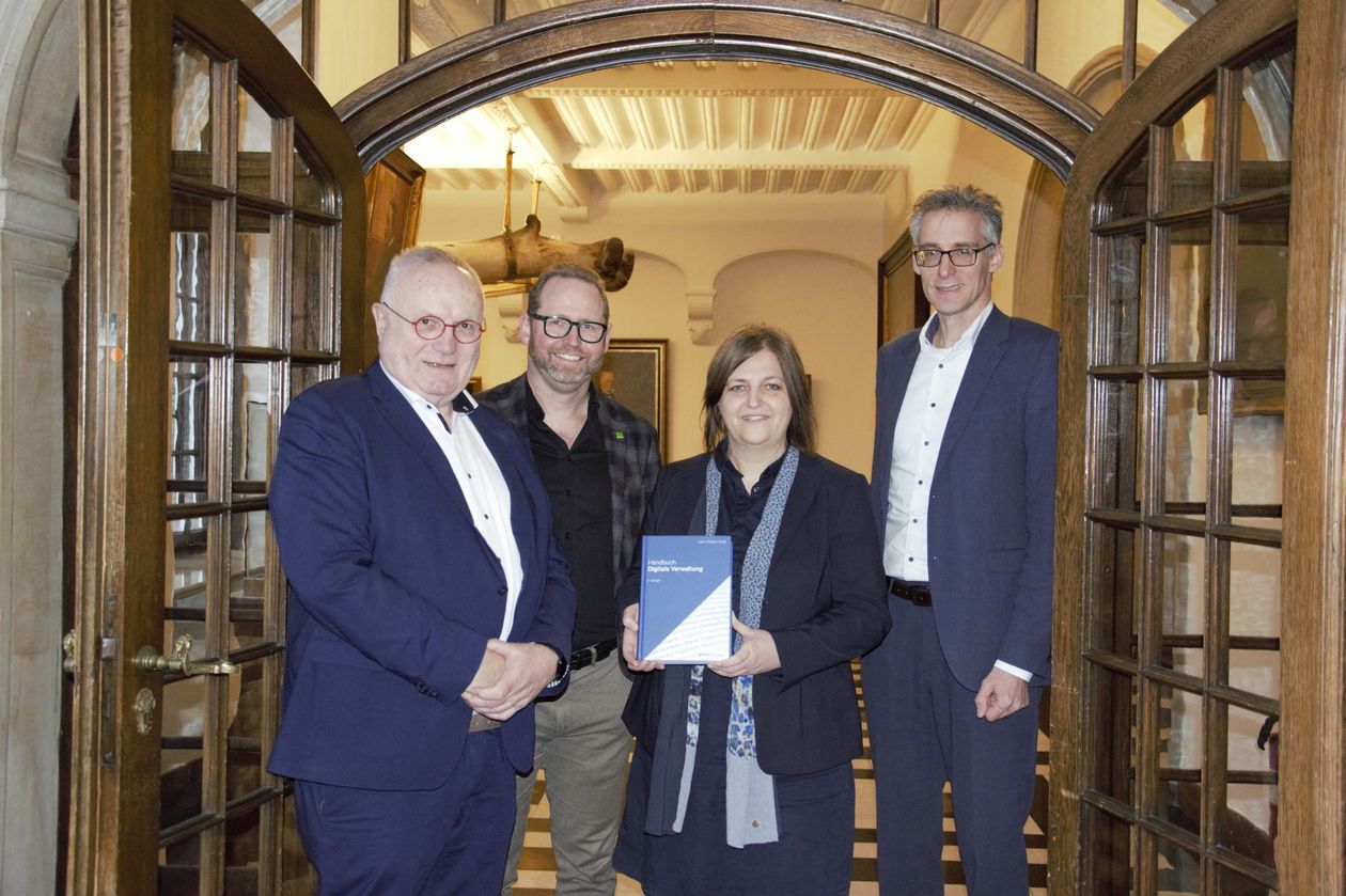 Gruppenbild mit drei Männern und einer Frau. Diese präsentiert in ihren Händen ein Buch. Alle stehen im Durchgang einer repräsentativen Holztür mit Glasfenstern. Beide Türflügel sind weit geöffnet.