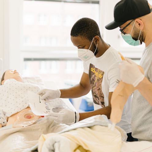 Pflegestudierende &uuml;ben an einem Krankenhausbett mit einer Puppe im Simulationszentrum des Studiengangs.