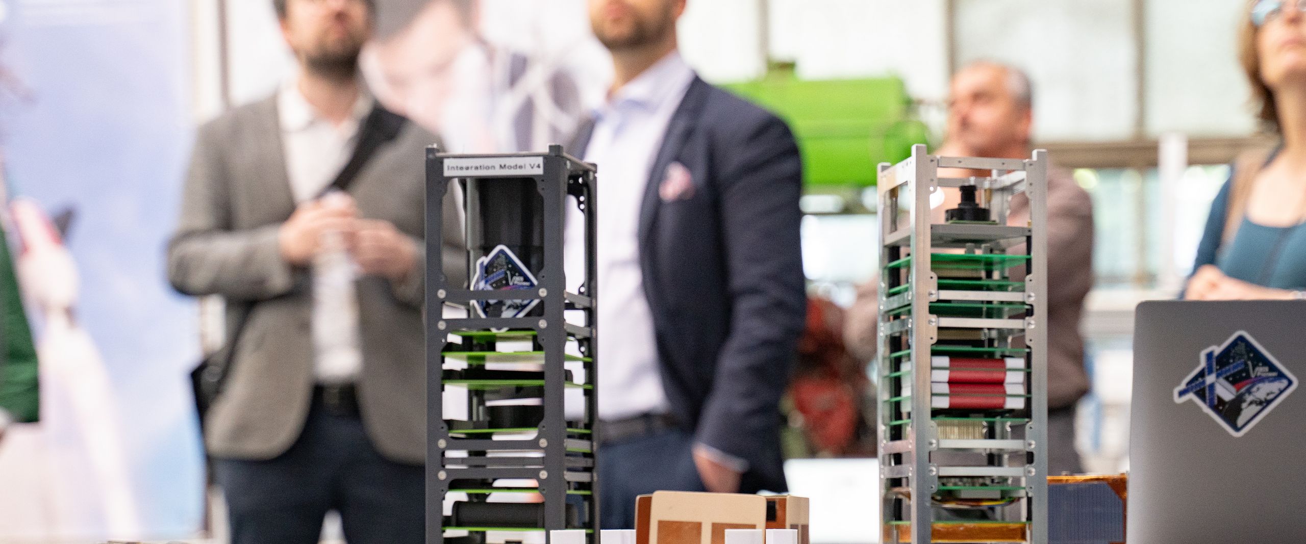 Auf dem Bild sind im Vordergrund zwei Prototypen eines Satelliten zusehen. Im Hintergrund sind unscharf vier Personen zu erkennen. 