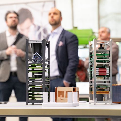 Auf dem Bild sind im Vordergrund zwei Prototypen eines Satelliten zusehen. Im Hintergrund sind unscharf vier Personen zu erkennen. 