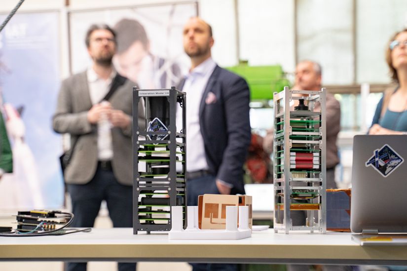 Auf dem Bild sind im Vordergrund zwei Prototypen eines Satelliten zusehen. Im Hintergrund sind unscharf vier Personen zu erkennen. 