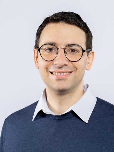 Auf dem Foto ist Karl Akbari zusehen. Er hat kurze schwarze Haare. Er tr&auml;gt eine runde Brille und ein wei&szlig;es Hemd unter einem blauen Pullover. 