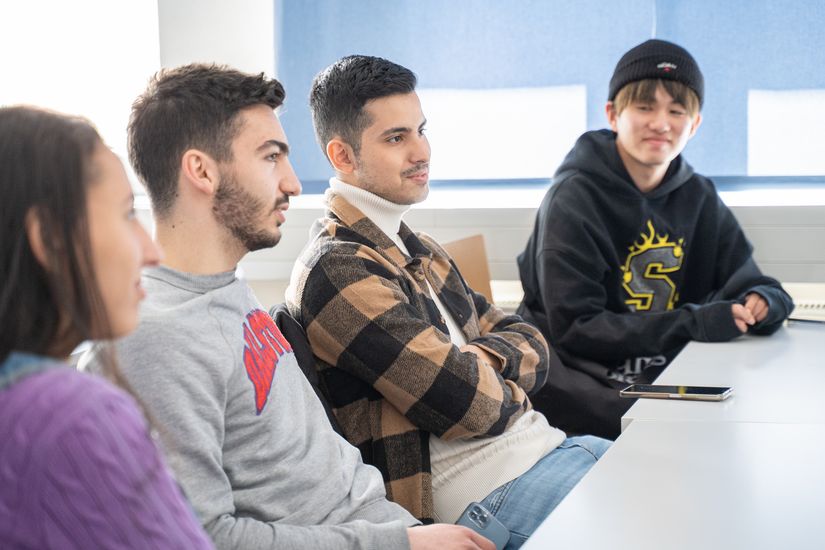 3 junge Studenten und eine Studentin sitzen an einem langen Tisch und schauen nach recht