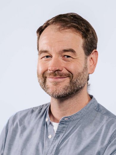 Auf dem Bild ist Hendrik Jungen zusehen. Er hat kurzes braunes Haar und einen Drei-Tage-Bart. Er trägt ein blau-graues Hemd mit Stehkragen. 