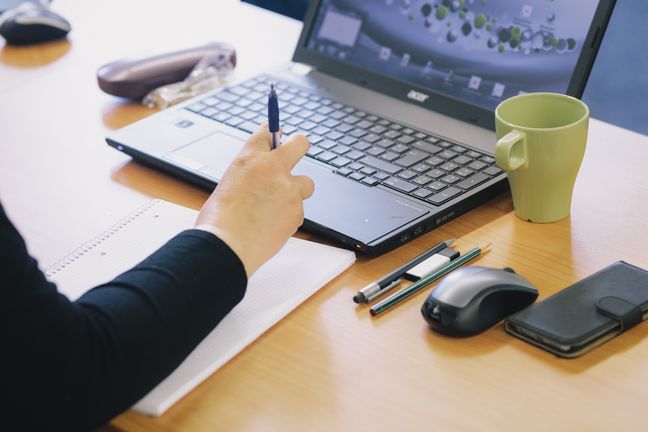 Schreiblabor  Arm auf einem Notizblock, Hand am Laptop, daneben Tasse, Stifte, Mouse und Handy