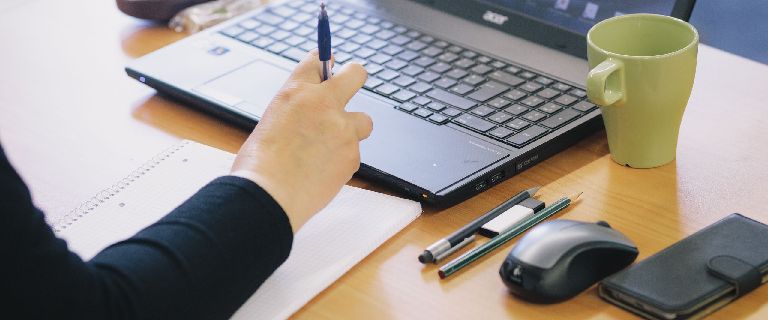 Arm auf einem Notizblock, Hand am Laptop, daneben Tasse, Stifte, Mouse und Handy