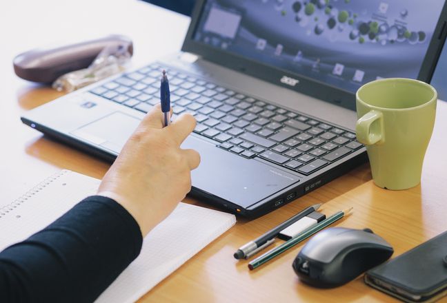 Schreiblabor  Arm auf einem Notizblock, Hand am Laptop, daneben Tasse, Stifte, Mouse und Handy